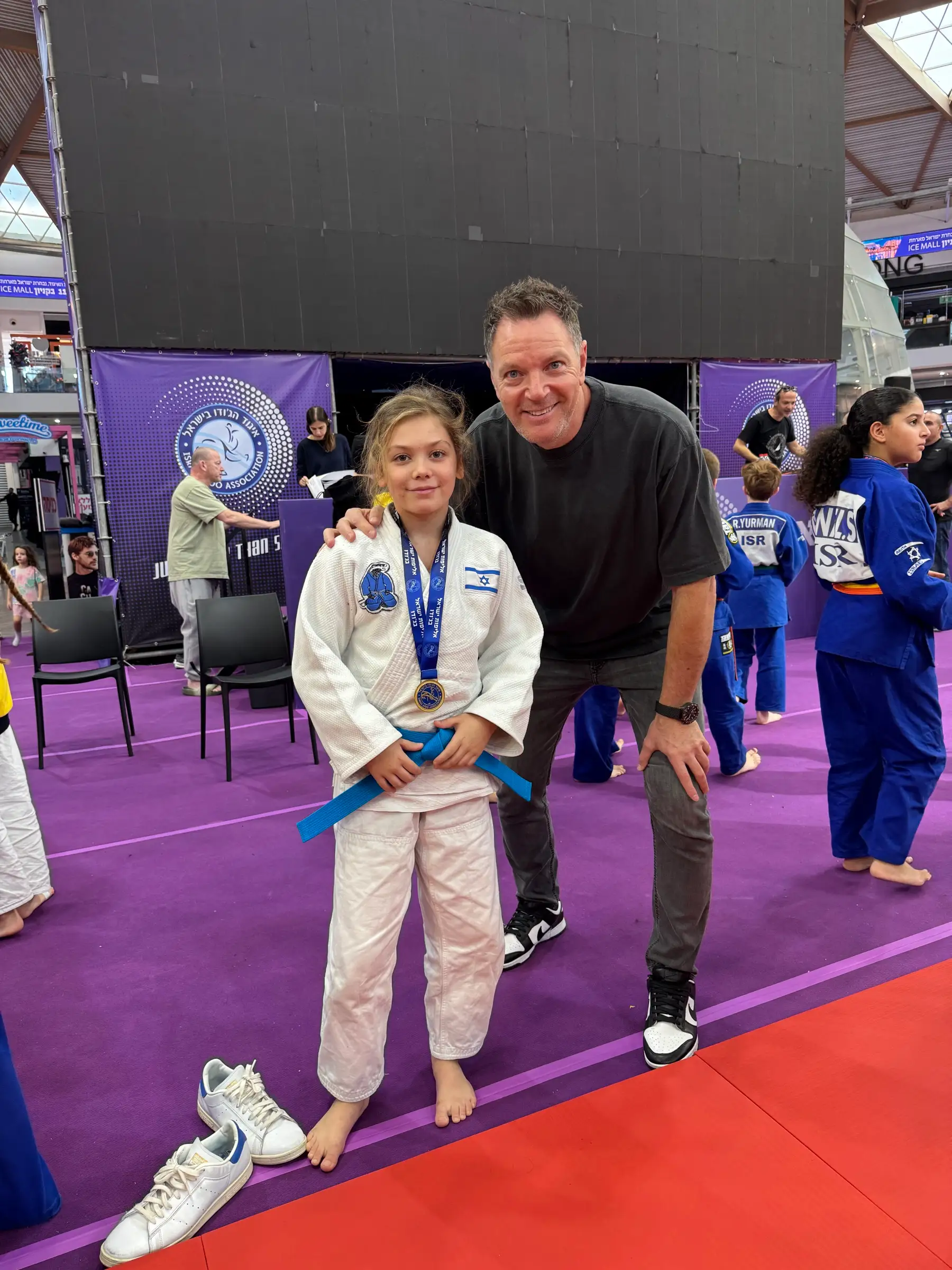 Melissa in a judo suit next to a man in black clothes, on a purple mat