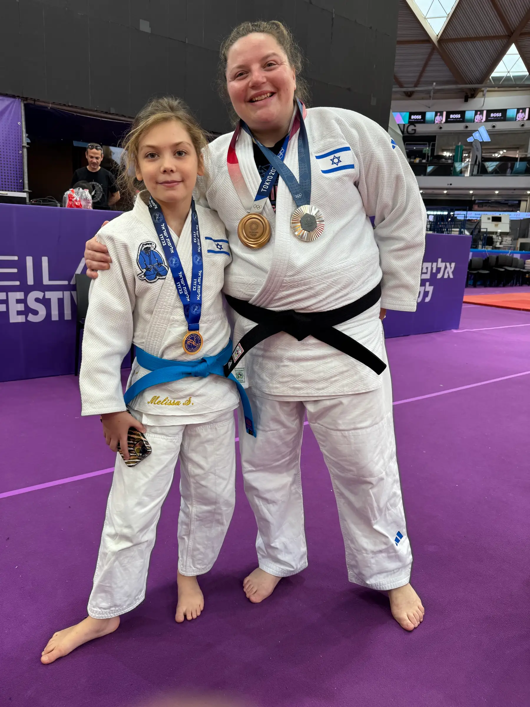Melissa and a woman in judo suits with medals and a black belt, on a purple mat