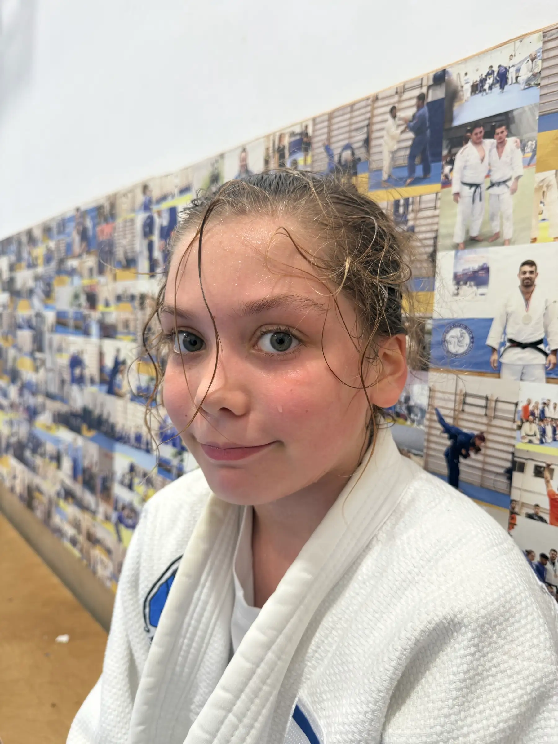 Melissa in a judo suit with wet hair after training, smiling in front of a wall of photos